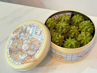 Decorative tin with green pastries and illustrated lid on a white surface